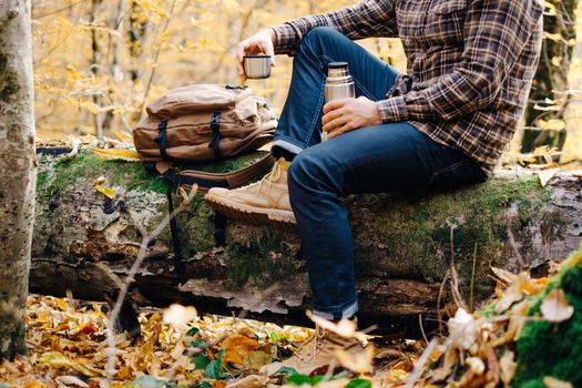 Unrecognizable young man resting with cup of tea in autumn forest.