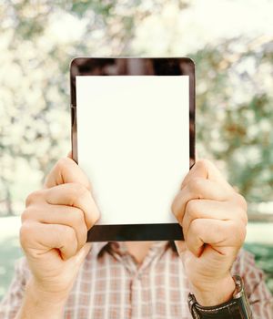 Young man holding tablet pc with empty screen in front of his face in summer park, close-up.