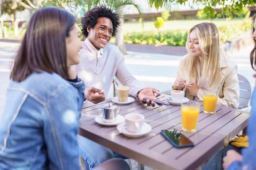 Multi-ethnic group of friends having a drink together in an outdoor bar. One of the men shows something on his smartphone to his friends.