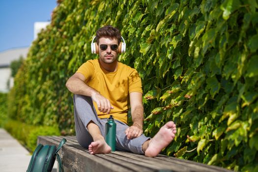 Male student sitting outside using an aluminum water bottle, headphones and backpack.