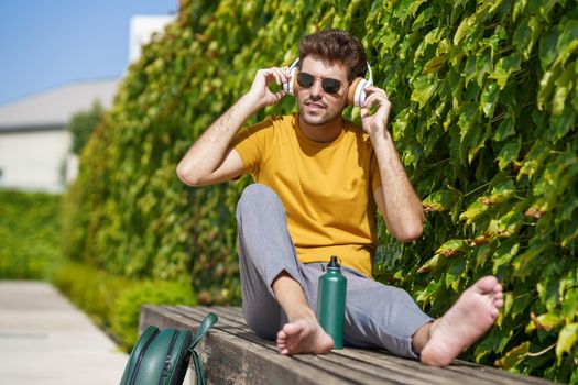 Male student sitting outside using an aluminum water bottle, headphones and backpack.