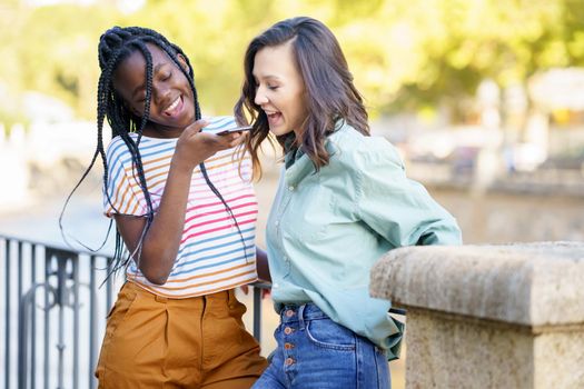 Two female friends recording a voice note with smartphone outdoors. Multiethnic friends.