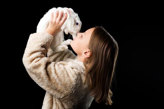Pretty stylish girl with white rabbit. Side shot of beautiful girl in beige fur coat looking at cute bunny pet. Portrait of preteen child posing in studio against black background