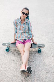 Smiling girl in sunglasses sitting on longboard, looking at camera.