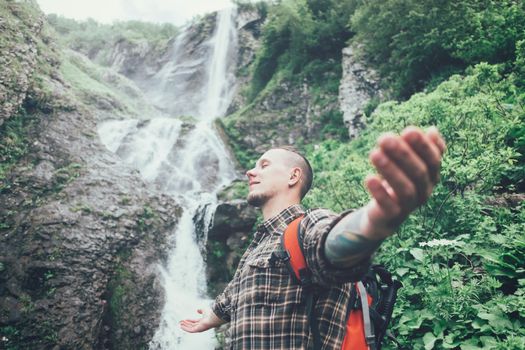 Happy backpacker young man standing with raised arms on background of waterfall and enjoying of summer nature, side view.