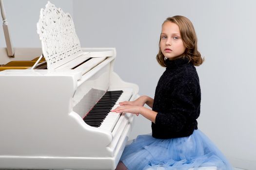 Beautiful teenage girl playing grand piano. Pretty blonde girl in stylish clothes learning to play music instrument. Teenage child looking at camera against gray background