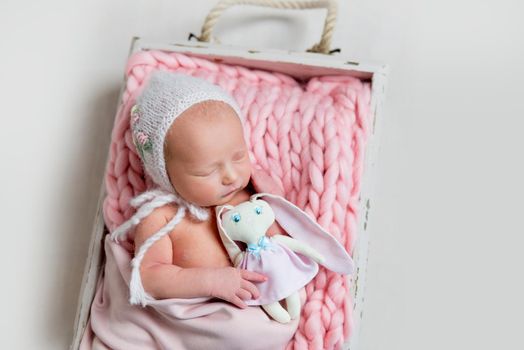 Sweet newborn girl in bonnet hat with chin strap sleeping and embracing her toy. Little girl sleeping on pink rug in wooden box, top view