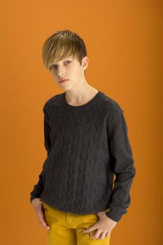 Stylish blond handsome teenage boy. Attractive boy wearing gray pullover and pants posing against ochre background. Portrait of stylish cheerful student teenager