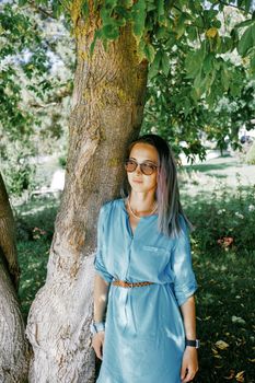 Stylish beautiful young woman wearing in blue dress and sunglasses standing near a tree in summer park.