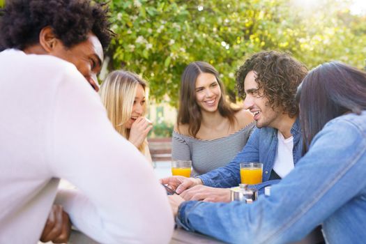 Multi-ethnic group of friends having a drink together in an outdoor bar. One of the men shows something on his smartphone to his friends.