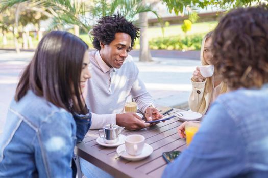 Black man showing something to his multi-ethnic group of friends on his smartphone, while having drinks at an outdoor table in a bar.