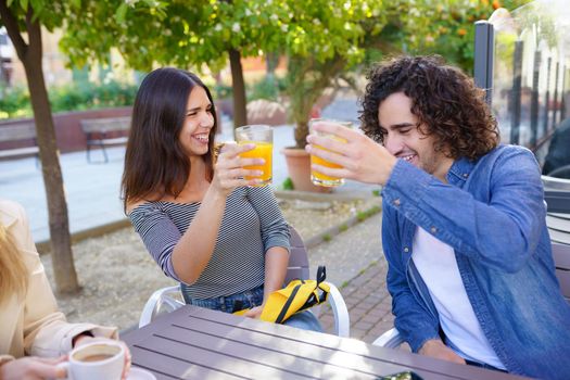 Couple of friends toasting with orange juices while having a drink with their multi-ethnic group of friends at the outdoor table of a bar.