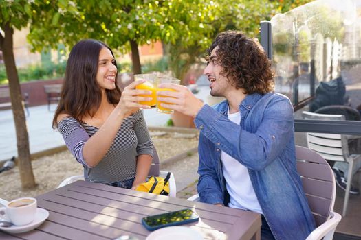 Couple of friends toasting with orange juices while having a drink with their multi-ethnic group of friends at the outdoor table of a bar.