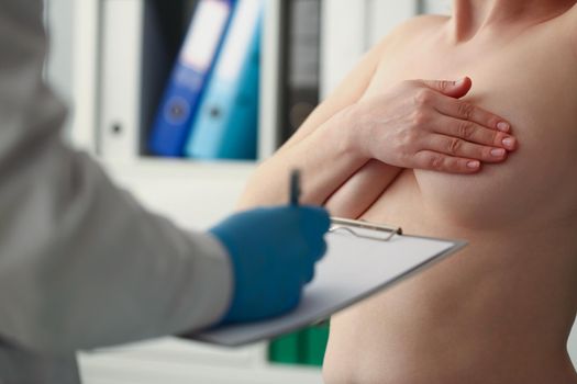 Close-up of woman close breast with hand, doctor make notes in prescription list. Female client wants breast implant, planned checkup at clinic before procedure. Plastic, cosmetic surgery concept