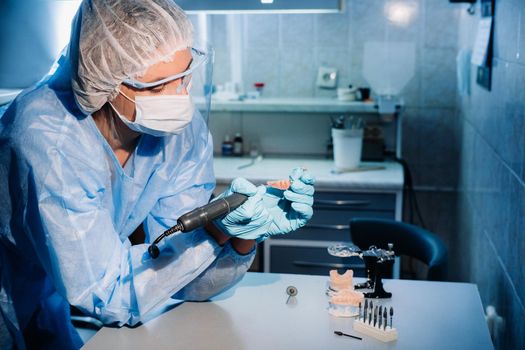 A masked and gloved dental technician works on a prosthetic tooth in his lab.