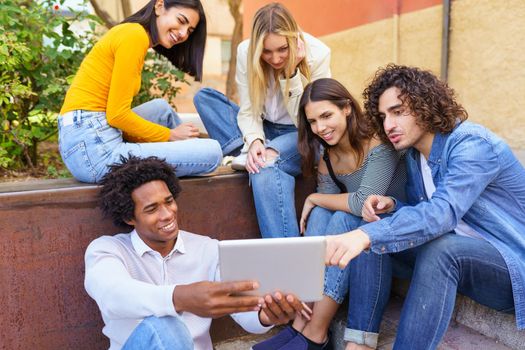 Multi-ethnic group of young people looking at a digital tablet outdoors in urban background. Group of men and woman sitting together on steps.