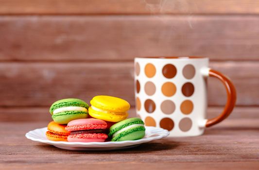 Sweet macarons dessert on a plate and cup of hot drink on wooden table.