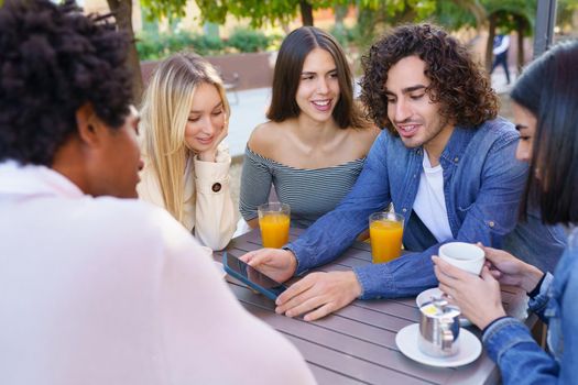 Young man showing something to his multi-ethnic group of friends on his smartphone, while having drinks at an outdoor table in a bar.