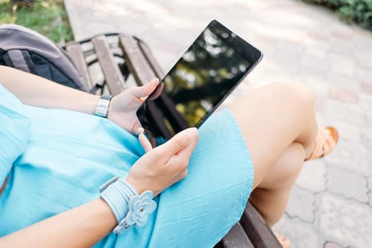 Unrecognizable young woman sitting with digital tablet on wooden bench in the park.