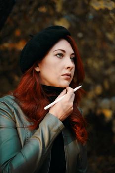 Beautiful red-haired woman in the park. Brooding woman in a beret and leather jacket. Man with a pen, designer, freelancer. Autumn portrait. Waiting for inspiration.