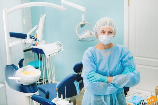 a female dentist wearing a medical mask and rubber gloves poses for the camera and folds her arms in her office.