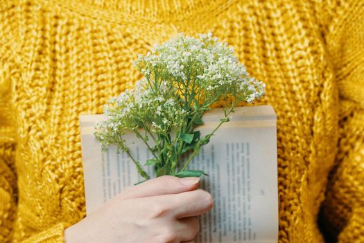 Bouquet in woman's hand on background of open book and knitted sweater. Close up. Girl with book and wildflowers. Romantic concept.