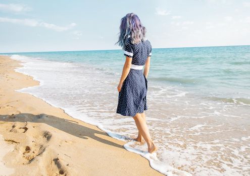 Unrecognizable barefoot young woman with blue hair in dress walking on shore near the sea.