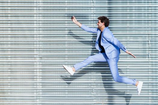 Young man wearing a suit makes a selfie with a smartphone while jumping outdoors