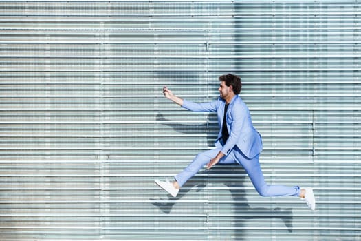 Young man wearing a suit makes a selfie with a smartphone while jumping outdoors