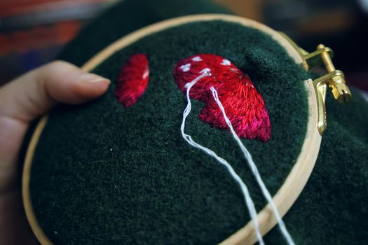 Girl's hand embroidery mushroom hat in hoop on green material. Concept of needlework, hobby, leisure. Close-up.