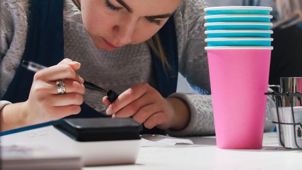 A woman barista writing something on the paper. Colorful cups on the foreground. Close up