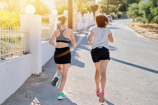 Running mother and teenage daughter, back view, along the street of a sunny summer city. Active healthy lifestyle, family and sports