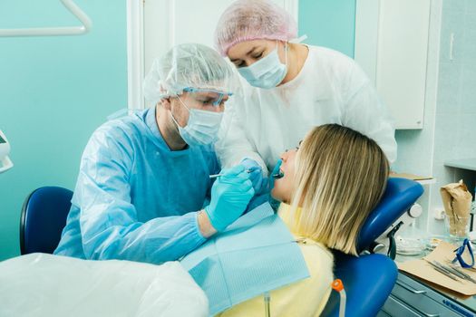 a dentist in a protective mask sits next to him and treats a patient in the dental office with an assistant.
