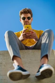 Young man using his smartphone sitting on a ledge outside. Guy wearing sunglasses.