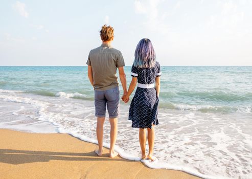 Young couple in love standing on sand beach and holding hands, looking at sea on travel summer vacations, rear view.