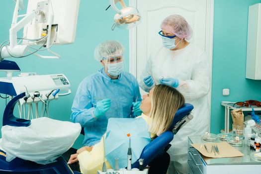 a dentist in a protective mask sits next to him and treats a patient in the dental office with an assistant.