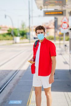 Student male wearing a surgical mask while waiting for a train at an outside station