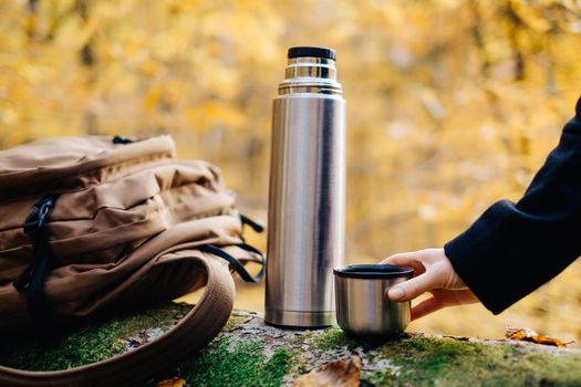 Female hand holding cup near the thermos in autumn forest.