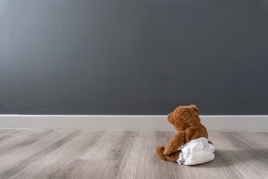 Brown bear in diaper sits near grey wall, good background, view from the back,enough space for text, concept of loneliness and sadness