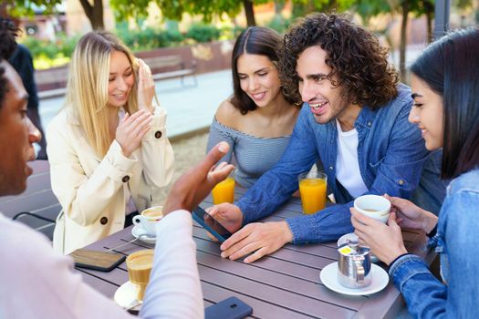 Multi-ethnic group of friends having a drink together in an outdoor bar. One of the men shows something on his smartphone to his friends.