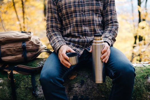 Unrecognizable young man resting in forest and holding cup and thermos.