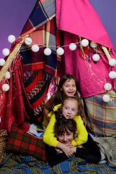 Kids embrace in home-made tent inside the living room before going to sleep. Child pajama party. Pajamas for kids. Little Girls in tipi house. Sisters or best friends spend time