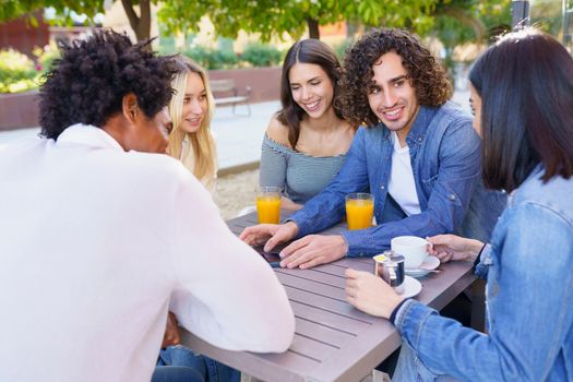 Young man showing something to his multi-ethnic group of friends on his smartphone, while having drinks at an outdoor table in a bar.