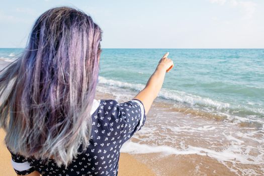 Unrecognizable young woman standing on sand beach and pointing at the sea, copy-space.