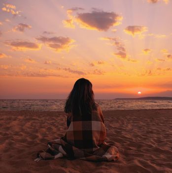 Young woman wrapped in plaid sitting on sand beach and enjoying view of sunset over sea.