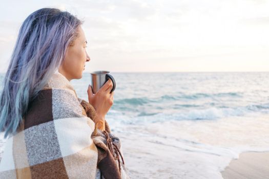 Smiling young woman wrapped in plaid resting with cup of beverage on beach near the sea.