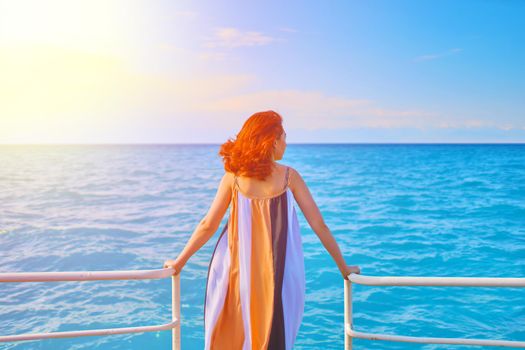 Cute girl in flowing long dress stands on edge of sea pier. Pretty woman is enjoying vacation. Red-haired beautiful woman looks at ocean. Endless water and blue sky. Hot summer day.