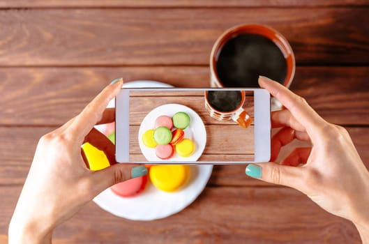 Woman taking photo of macarons dessert with cup of coffee with smartphone, top view of hands.