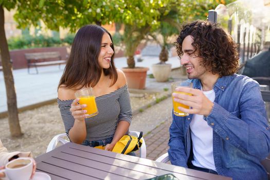 Couple of friends toasting with orange juices while having a drink with their multi-ethnic group of friends at the outdoor table of a bar.