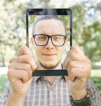Young man holding digital tablet with smiling male portrait in front of his face in summer park.